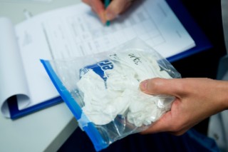 Eine Hand einer Person hält einen Plastikbeutel mit benutzten Papiertaschentüchern; die andere Hand notiert etwas auf einem Klemmbrett.