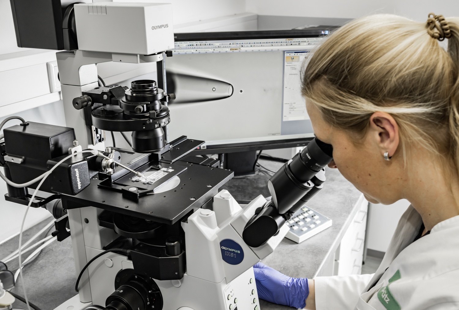 Wissenschaftlerin am Mikroskop; im Hintergrund ein Bildschirm mit einer Pipette und einer einzelnen Zelle.