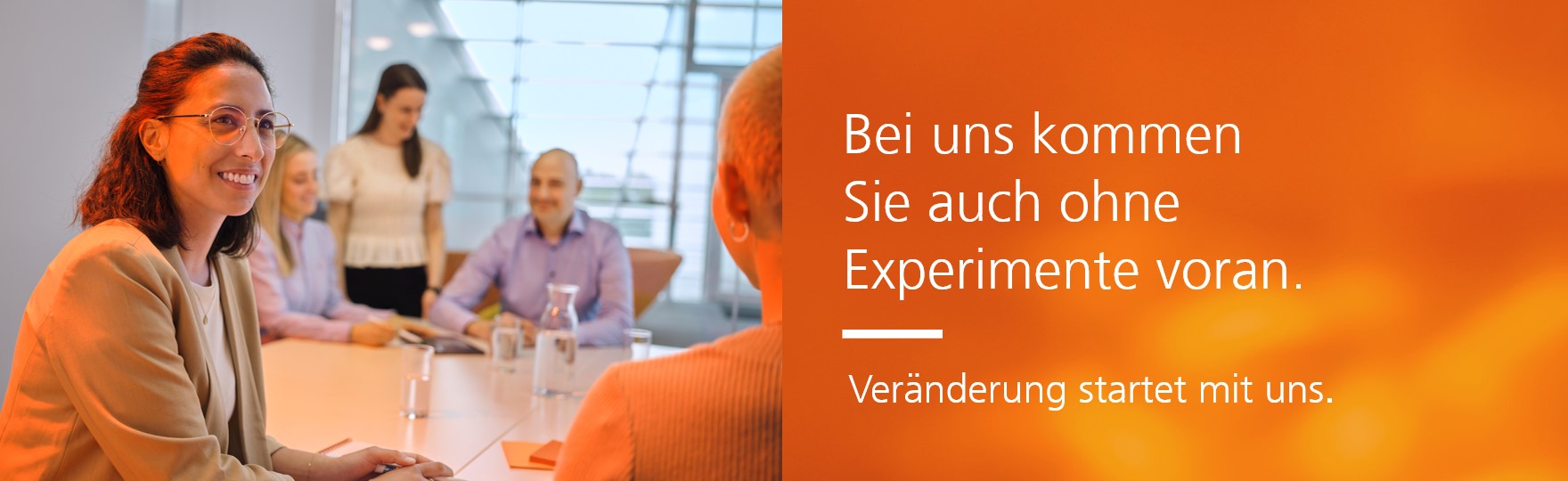 Eine Gruppe von fünf Personen sitzt in einem Konferenzraum an einem Tisch, eine Frau im Vordergrund trägt eine Brille und lächelt, im Hintergrund sitzen und stehen weitere Personen.  Daneben der Spruch "Bei uns kommen Sie auch ohne Experimente voran. Veränderung startet mit uns"