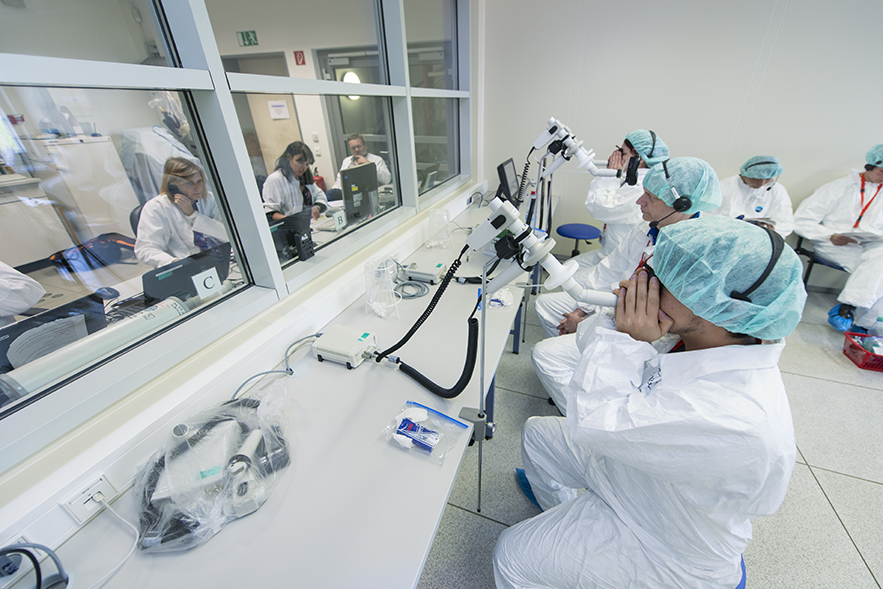 View into a pollen exposure chamber with test subjects, monitored by medical staff behind a glass panel.