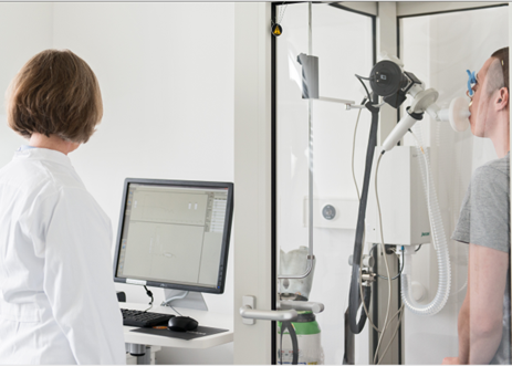 Test subject undergoing a lung function test in a chamber; a medical assistant looks at a PC screen.